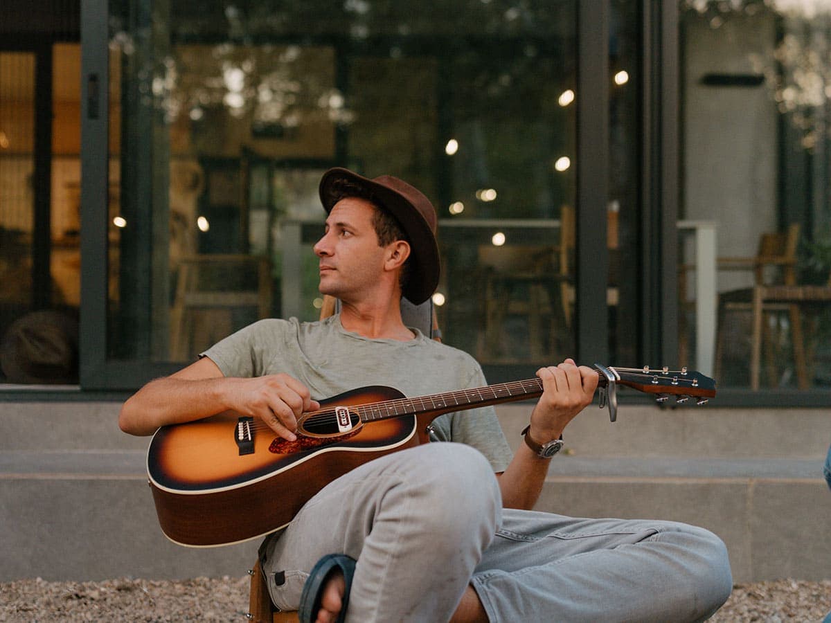 A man sits on a bench, strumming an acoustic guitar, enjoying a moment of music in a serene outdoor setting